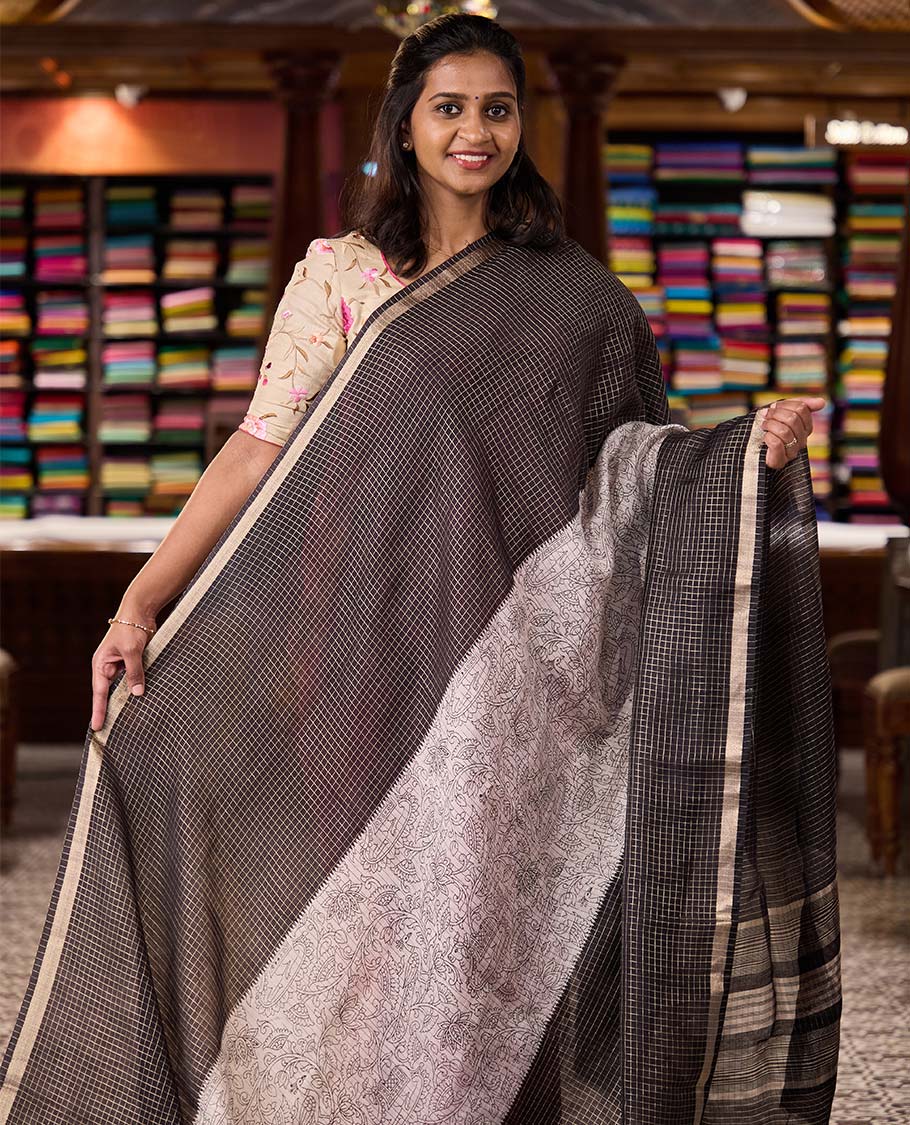 Black and beige half-and-half style Muga silk saree adorned with zari micro-checks, and kalamkari flora-fauna prints, zari piping border, and zari striped pallu