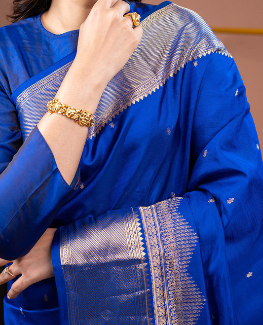 Blue Vasundhara Silk Saree Gleamed with Zari Buttas All Over Its Body, Highlighted with a Zari Border of Thilakam Mokku, Muthu, Cross Kamalam, and Mayil Kann Designs, Grand Pallu of Jaal Design, and Paired with a Matching Blouse